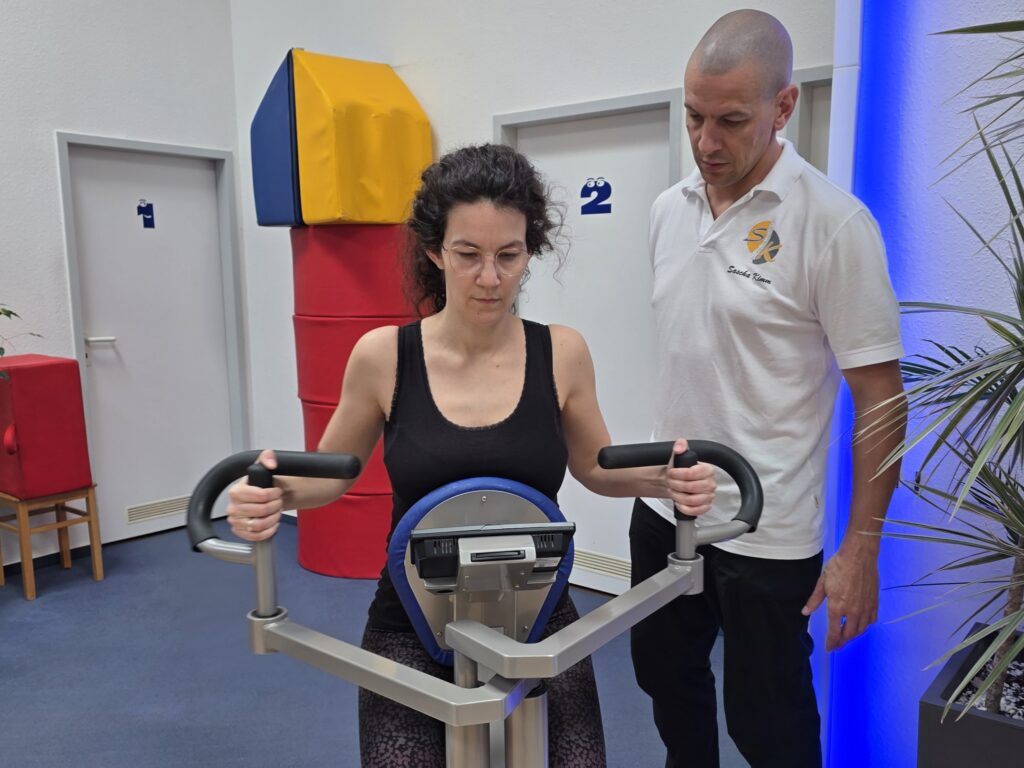 Frau trainiert an einem Fitnessgerät, Therapeut steht daneben und gibt Anleitung.