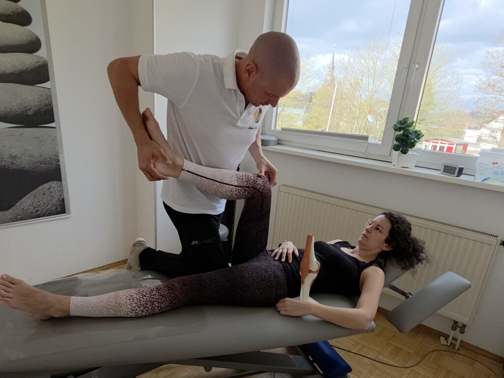 Therapeut bewegt das Bein einer liegenden Patientin zur Mobilisation im Behandlungsraum.