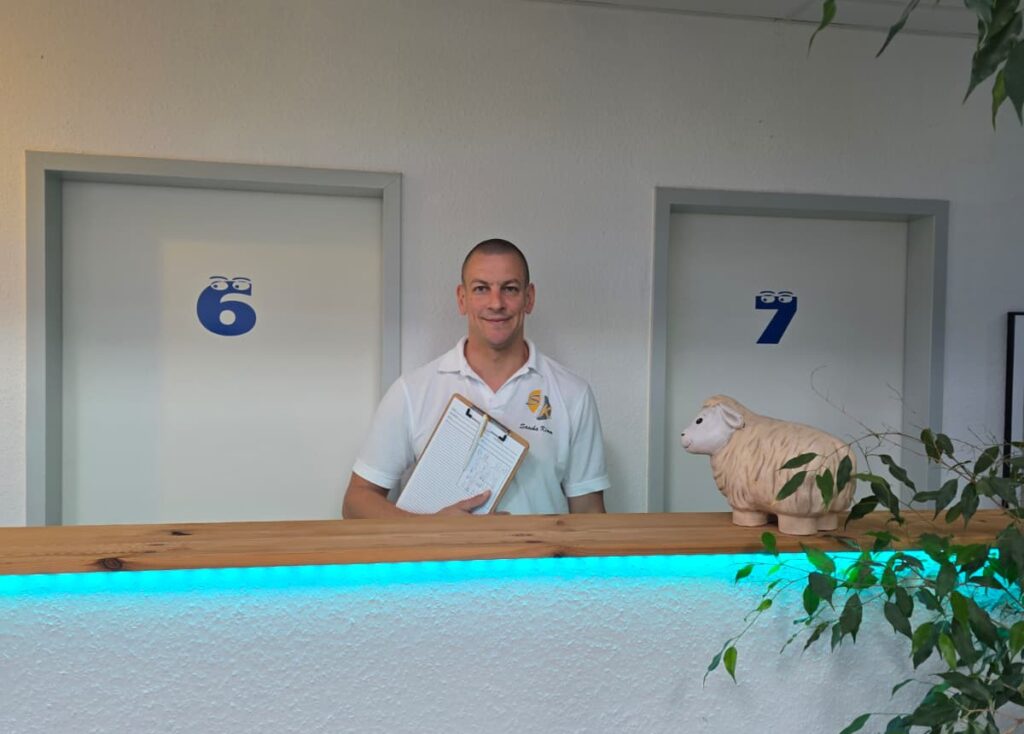 Mitarbeiter steht am Empfang mit Klemmbrett vor Behandlungsräumen und blickt freundlich nach vorn.